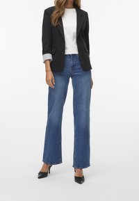 Svart kavaj med slag över en vit t-shirt, kombinerat med ljusblå vida jeans och svarta spetsiga klackskor. Slät tygkvalitet och skräddarsydd passform.