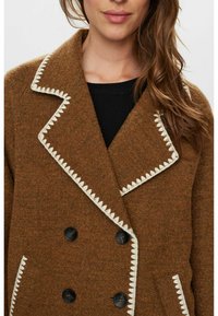 Manteau en mélange de laine brun avec un design à double boutonnage, une couture contrastante en blanc en forme de feston le long du col et des boutons noirs.