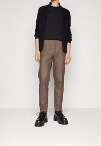 Svart zip-up cardigan, åtsittande svart t-shirt, bruna texturerade byxor och svarta skor med tjocka sulor, stående mot en neutral bakgrund.