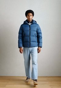 Geacă puffer de culoare albastră închisă, cu fermoar în față, glugă și manșete elastice. Asortată cu blugi de culoare albastră deschisă și încălțăminte casual.