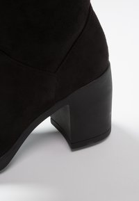 Bottine noire en daim avec un talon carré. Le talon est entièrement noir, offrant une transition harmonieuse avec la tige. Détail de surface texturé.