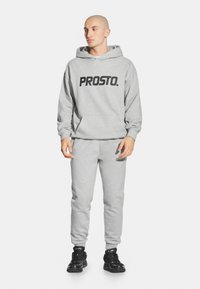 Szara bluza z kapturem z dużym czarnym logo "PROSTO.", w zestawie ze szarymi dresami. Materiał wydaje się być miękki; dopasowane mankiety i pas. Czarne sneakersy.