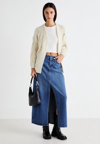 Cremfarbener Kabelstrick-Cardigan, weißes Cropped-Top, blaue Jeans-Maxiröhre mit einem vorderen Schlitz, schwarze Stiefeletten und schwarze Schultertasche.