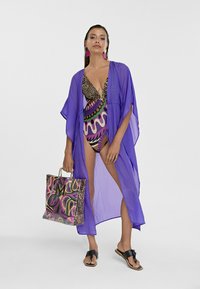 Kimono trasparente viola sopra un costume da bagno a stampa leopardata e colorata, abbinato a una borsa a tracolla con motivo e infradito neri.