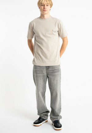 Młody mężczyzna stojący z rękami za plecami, ubrany w beżową koszulkę, luźne szare dżinsy i czarne trampki na białym tle.