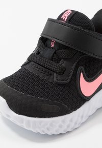 Schwarzer Sportschuh mit strukturiertem Mesh-Obermaterial, pinkem Nike-Swoosh, Klettverschluss und weißer Gummisohle mit Blasenmuster.