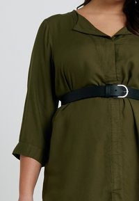 Indumento verde oliva a maniche lunghe con collo, vita cinchata e cintura nera con fibbia argentata. Tessuto liscio, chiusura a bottoni davanti.