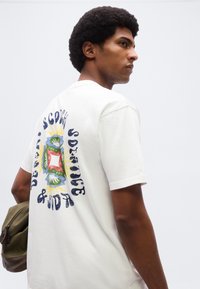 Άνδρας με χτένισμα αφρο, φοράει λευκό γραφικό μπλουζάκι και κρατάει σακίδιο πλάτης ελιάς, κοιτάζοντας προς τα δεξιά σε απλό φόντο.