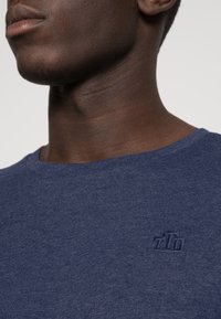 Mörkblå t-shirt med kort ärm som är tillverkad av textil med struktur, prydd med en diskret broderad logotyp på bröstet och en klassisk rund halsringning.