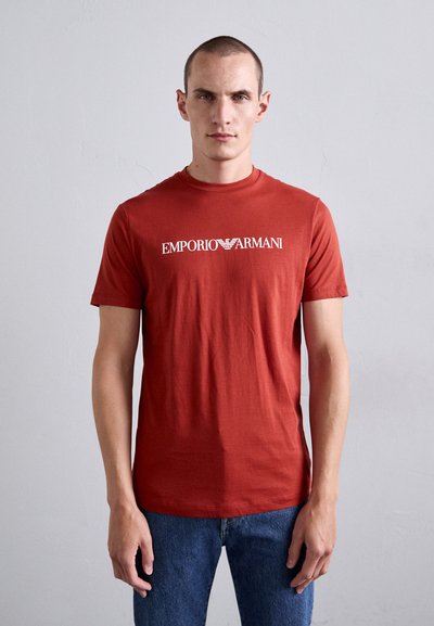 Mężczyzna z krótkimi włosami, ubrany w czerwoną koszulkę Emporio Armani i niebieskie dżinsy, stojący na jednolitym jasnoszarym tle.