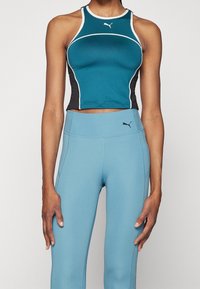 Donna che indossa un top sportivo senza maniche Puma teal e nero e leggings Puma a vita alta azzurri, in piedi davanti a uno sfondo semplice.