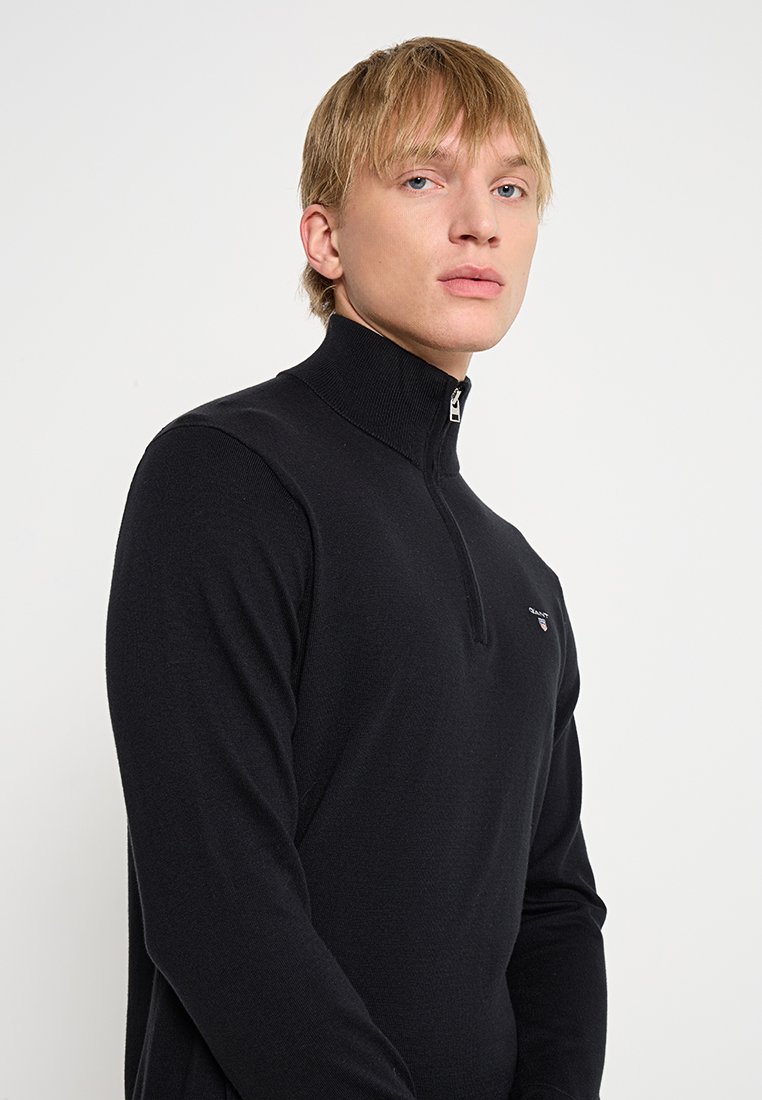 Uomo biondo con occhi chiari che indossa un maglione a zip corto nero con un piccolo logo sul petto, in piedi contro uno sfondo bianco semplice.