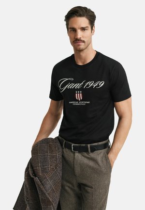 Zwarte t-shirt met korte mouwen met "Gant 1949" in witte schrift en een rood-wit-blauw schildlogo. Model draagt grijze gestructureerde broek en houdt een bruine jas vast.