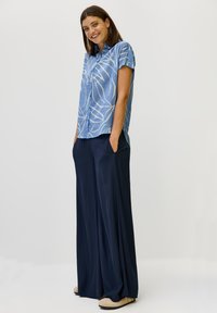 Camicia azzurra a maniche corte con motivi a foglia bianchi, abbinata a pantaloni larghi blu marino. Il modello è in piedi con le mani in tasca, indossando scarpe beige.