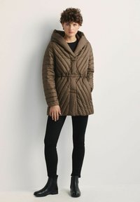 Brązowa pikowana kurtka puffer z dużym kapturem, ściągnięta w talii, z pionowymi szwami. Noszona na czarnych legginsach i botkach za kostkę.