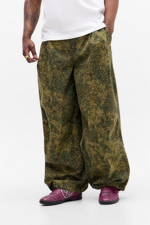 Man wearing loose digital camouflage pants, white t-shirt, purple sneakers, and a gold ring, standing with one hand in pocket.