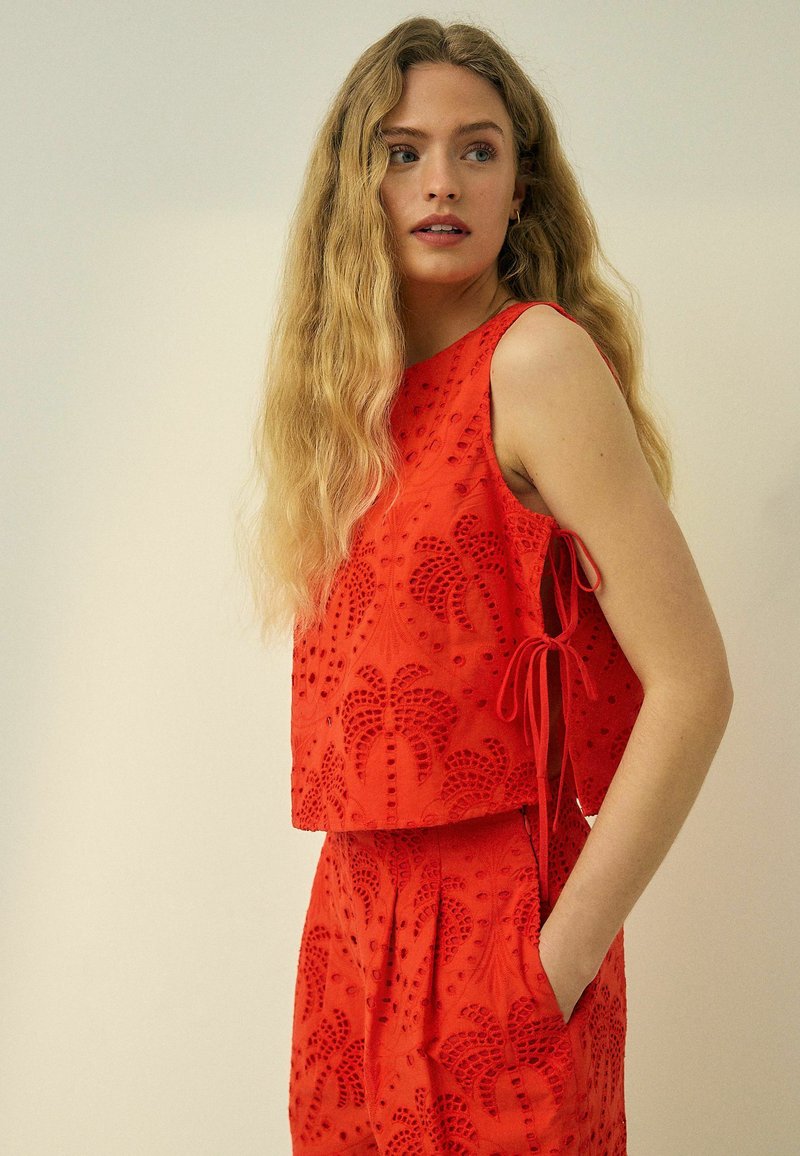 Femme aux longs cheveux blonds portant un haut rouge sans manches à œillets et un short taille haute assorti avec des poches latérales, regardant sur le côté.