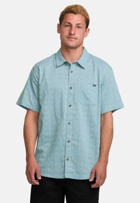 Camisa de manga corta en tejido azul claro, con un patrón de diamantes, cierre frontal con botones, cuello y un bolsillo en el pecho con un parche de logo.