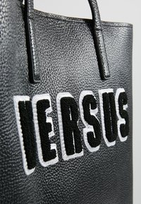 Sac cabas en cuir noir texturé avec de grandes lettres brodées en noir et blanc formant le mot "VERSUS" sur le devant.