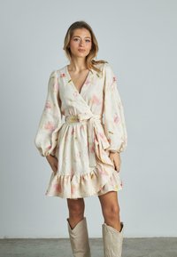 Wickelkleid aus leichtem Stoff mit floralem Muster in Pastellfarben. Es hat puffärmel und einen rüschensaum. Tragen mit beigen Stiefeln.