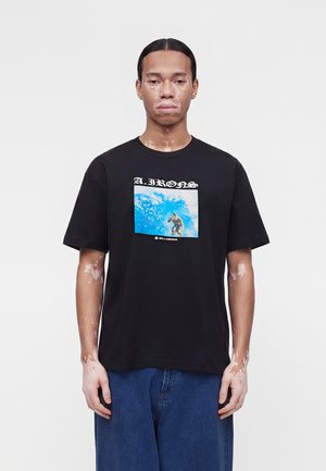 Persoană cu vitiligo stă cu fața spre înainte, purtând un tricou negru cu o grafică albastră de surfing și blugi largi albaștri pe un fundal simplu.