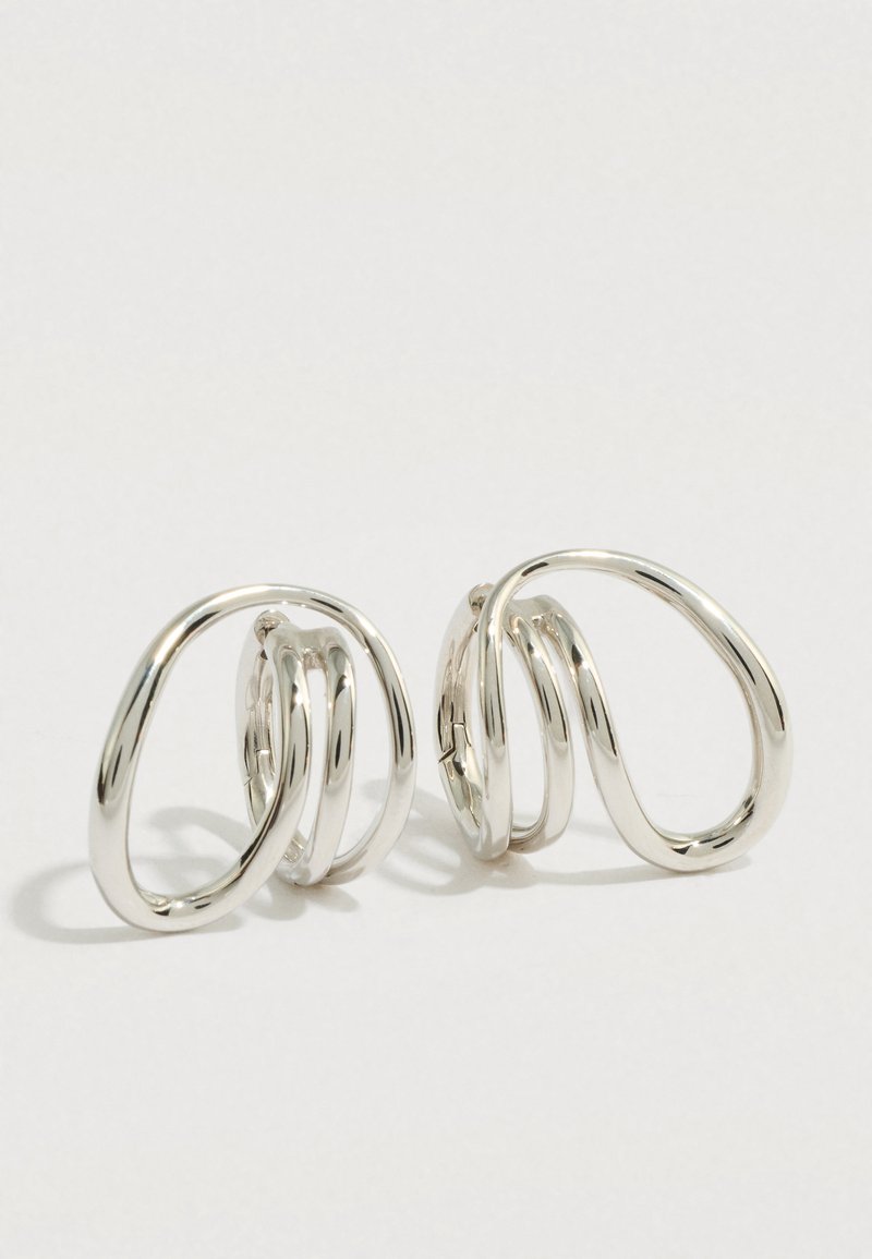 Ζευγάρι λαμπερά ασημένια σκουλαρί κια με αφηρημένα, ανοιχτά σχέδια σε σχήμα κύκλου σε απλό φωτεινό φόντο.