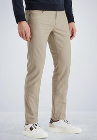 Chinos en mélange de coton beige avec une coupe slim, dotés de poches latérales, d'une texture distinctive et associés à des baskets blanches et noires.