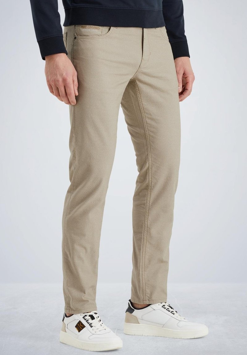 Chinos en mélange de coton beige avec une coupe slim, dotés de poches latérales, d'une texture distinctive et associés à des baskets blanches et noires.