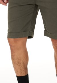 Olivgröna bomullsshorts med uppvikta nederkanter, slät textur och rak passform, delvis avslöjande av ett ben och en svart socka.