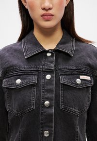 Jaqueta de ganga preta com colarinho clássico, botões de pressão prateados e dois bolsos no peito com um pequeno logo da Calvin Klein.