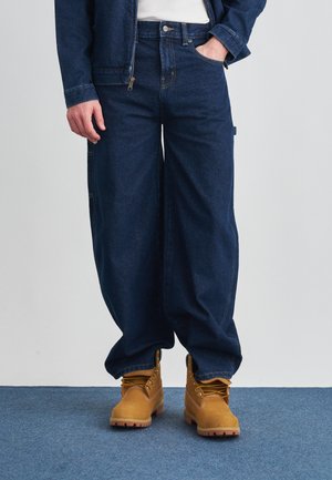 Pantaloni di denim blu scuro con vestibilità ampia, dotati di grandi tasche laterali, abbinati a stivali color cuoio con lacci e dettagli gialli.