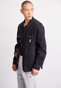 Black denim shirt with two chest pockets and snap buttons, paired with gray sweatpants featuring a logo. A small crossbody bag is worn.