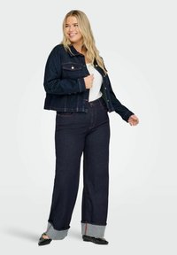 Kvinna som ler, iklädd en mörk denimjacka och vida jeans med uppvikta fållar, vit topp och svarta spetsiga ballerinas, stående framför vit bakgrund.