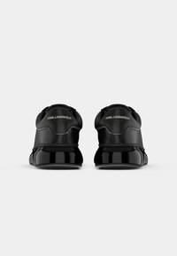 Ténis pretos com um acabamento liso e brilhante, apresentando um design minimalista e uma marca na parte de trás. Com calcanhar arredondado e suporte acolchoado para o tornozelo.