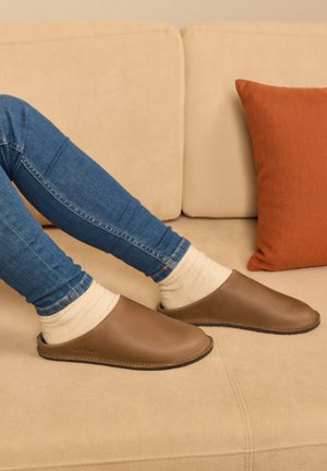 Beine in blauen Jeans und cremefarbenen Socken, die braune Leder-Hausschuhe tragen und auf einem beigen Sofa mit orangem Kissen ruhen.
