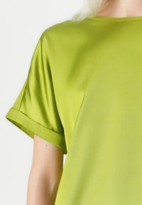 Short-sleeve top in bright green satin fabric, featuring a simple round neckline and rolled cuffs, smooth texture with no visible patterns.