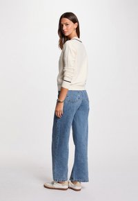 Frau mit langen braunen Haaren, die einen cremfarbenen Pullover, blaue Jeans, weiße Turnschuhe und goldene Armbänder trägt, steht vor einem schlichten weißen Hintergrund.