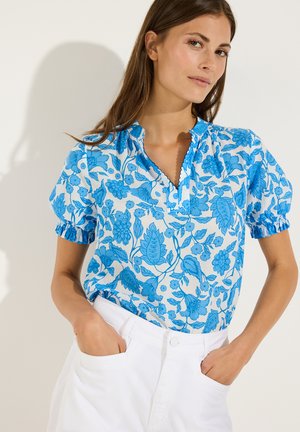 Femme portant un chemisier à manches courtes bleu et blanc à motif floral avec col à volants et pantalon blanc taille haute, mains dans les poches.