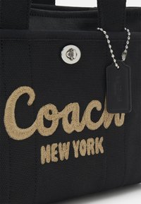 Sac fourre-tout en tissu noir avec broderie beige "Coach New York", fermeture à tourniquet argentée, et étiquette en cuir noir Coach attachée par une chaîne à billes.
