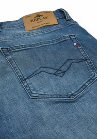 Jeans de mezclilla con un lavado azul claro, que presentan una etiqueta de cuero marrón y un bordado intrincado en el bolsillo trasero. Diseño clásico de cinco bolsillos.