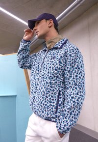 Jachetă din fleece cu fermoar, de culoare albastru deschis, cu pete de leopard în albastru închis și teal; are guler înalt și buzunare frontale cu fermoar.