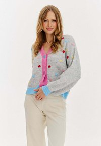 Szary wełniany kardigan z kolorowymi kropkami, różowymi guzikami z przodu oraz niebieskimi ściągaczami przy mankietach i u dołu. Miękka faktura i luźny krój.