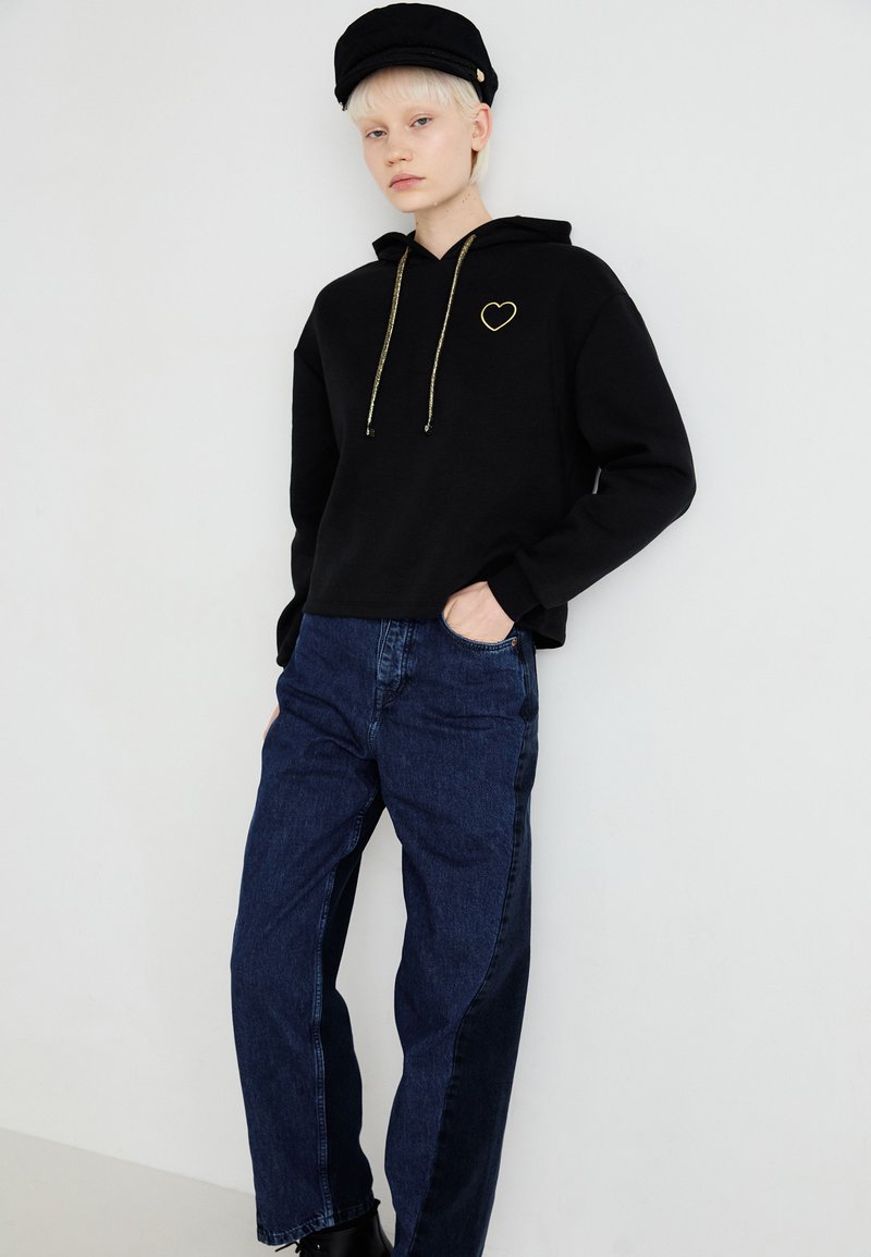 Felpa nera con cappuccio e logo a forma di cuore dorato, abbinata a jeans blu scuro. La felpa ha una vestibilità comoda e dettagli con lacci dorati.