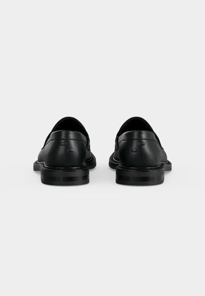 Paire de chaussures habillées en cuir noir vues de dos avec des petits talons empilés sur un fond gris clair uni.