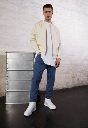 Un hoodie zippé crème clair, un t-shirt blanc, un jean en denim bleu et des baskets montantes blanches ; se tenant à côté d'une benne de stockage métallique.