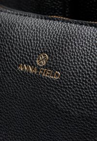 Svart läderväska med en strukturerad yta. Har en guld "ANNA FIELD"-logotyp. Närbild visar fin detaljering och hög kvalitet på hantverket.