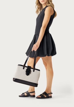 Donna che indossa un abito nero senza maniche e sandali neri, che tiene in mano una borsa shopper beige e nera per i manici, in piedi davanti a uno sfondo bianco.