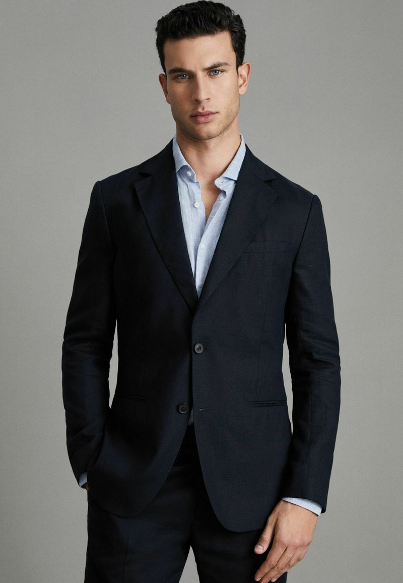 Blazer bleu marin cu un croi ajustat, material texturat, două butoane, rever în formă de notă și o cămașă cu guler de culoare albastru deschis dedesubt.