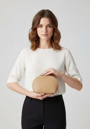 Pochette ovale beige texturée avec une fermeture éclair, tenue dans des mains. Un haut blanc uni et un pantalon noir créent un contraste avec la couleur du produit.