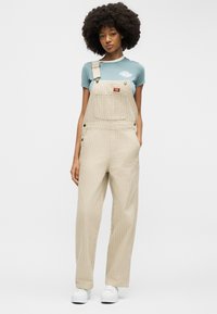 Jeune femme afro portant un t-shirt bleu clair, une salopette beige rayée avec une bretelle détachée, et des baskets blanches, debout sur fond blanc.